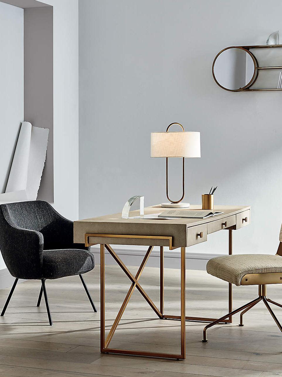 On a gold-framed desk between two dark chairs, this Clérachic table lamp sits lit in a bright, minimal home office setting.
