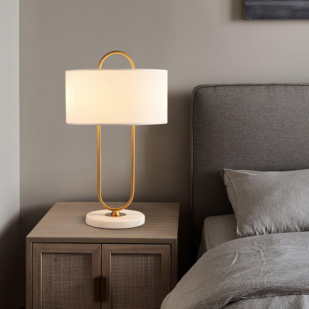 A Clérachic table lamp with its brass loop and white drum shade stands on a light wood nightstand beside a grey upholstered headboard.