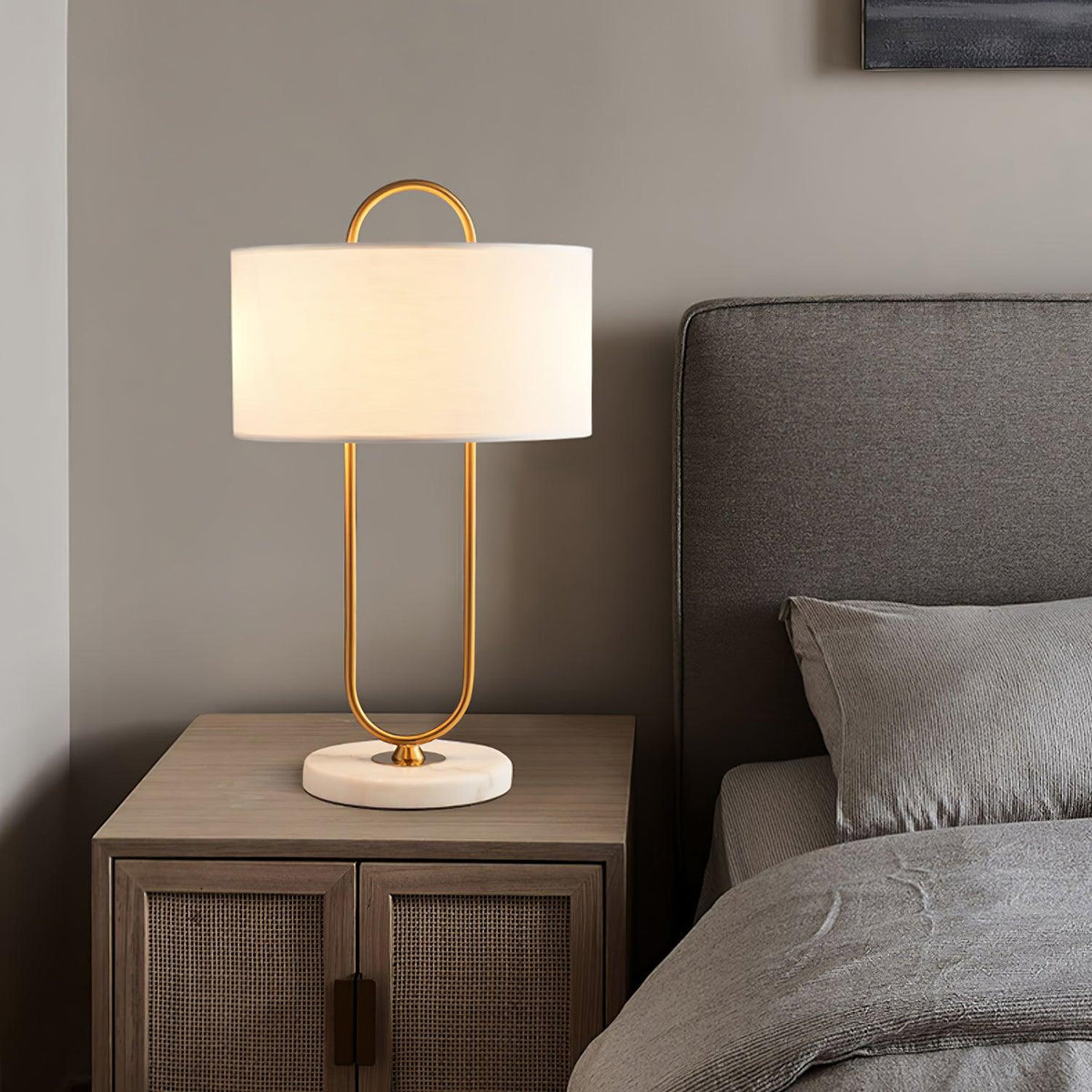 A Clérachic table lamp with its brass loop and white drum shade stands on a light wood nightstand beside a grey upholstered headboard.
