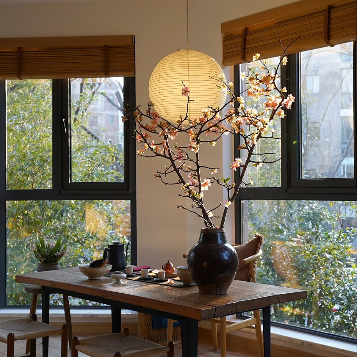 Flowering branches sit on a rustic table beneath a lit mulberry paper globe from Clérachic near tall windows.