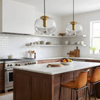 Two clear glass pendants with gold frames hang above a dark wood kitchen island with white countertops and leather bar stools, this Clérachic light shown in a pair.