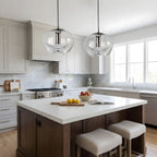 Two clear glass pendants with silver frames hang side by side above a white kitchen island with a marble backsplash, this Clérachic fixture shown in a pair.
