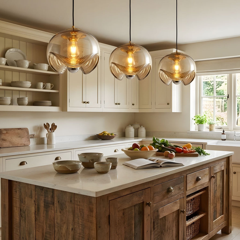 Three amber glass shades with gold frames hang in a row above a rustic wood-and-white kitchen island, this Clérachic fixture shown as a triple cluster.