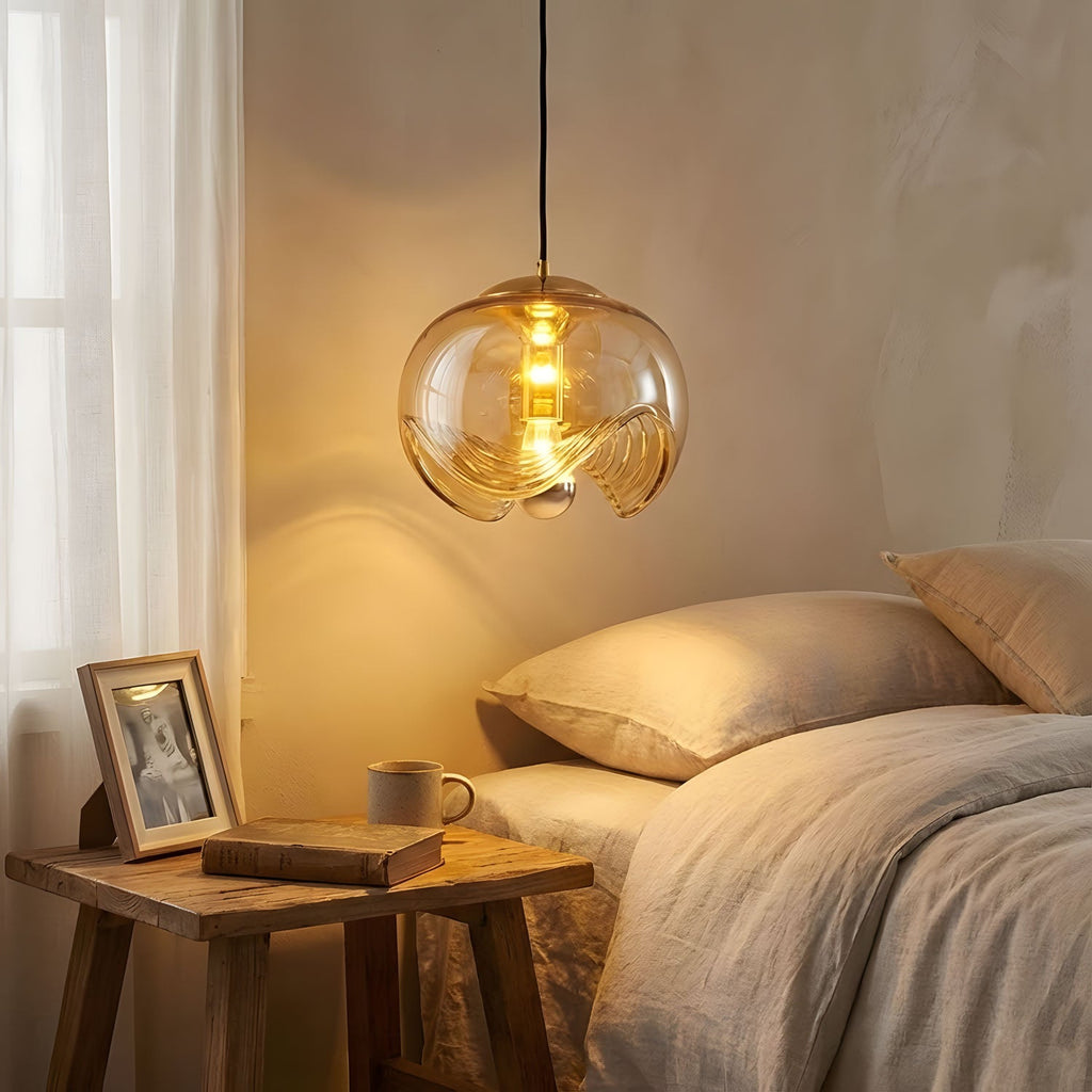 Lit warmly at dusk beside a nightstand stacked with books, this Clérachic pendant's amber glass shade glows above soft bedding.