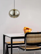 Above a white table set with cane-back chairs and a bowl of peaches, this Clérachic pendant's smoky grey glass shade and gold frame hangs against a plain wall.