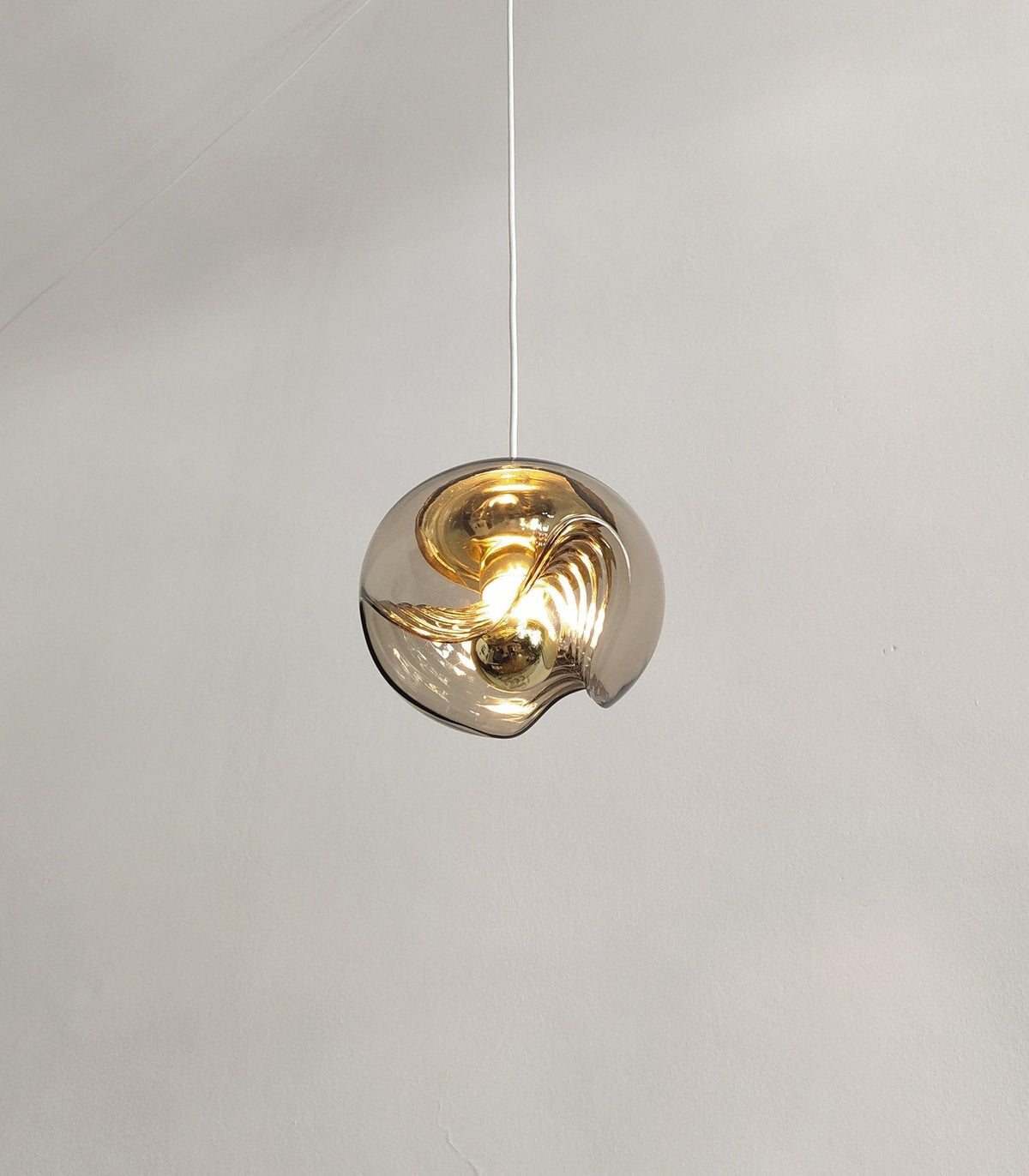 Viewed from below on a white cord, this Clérachic pendant's amber glass shade glows warmly against a plain grey background.