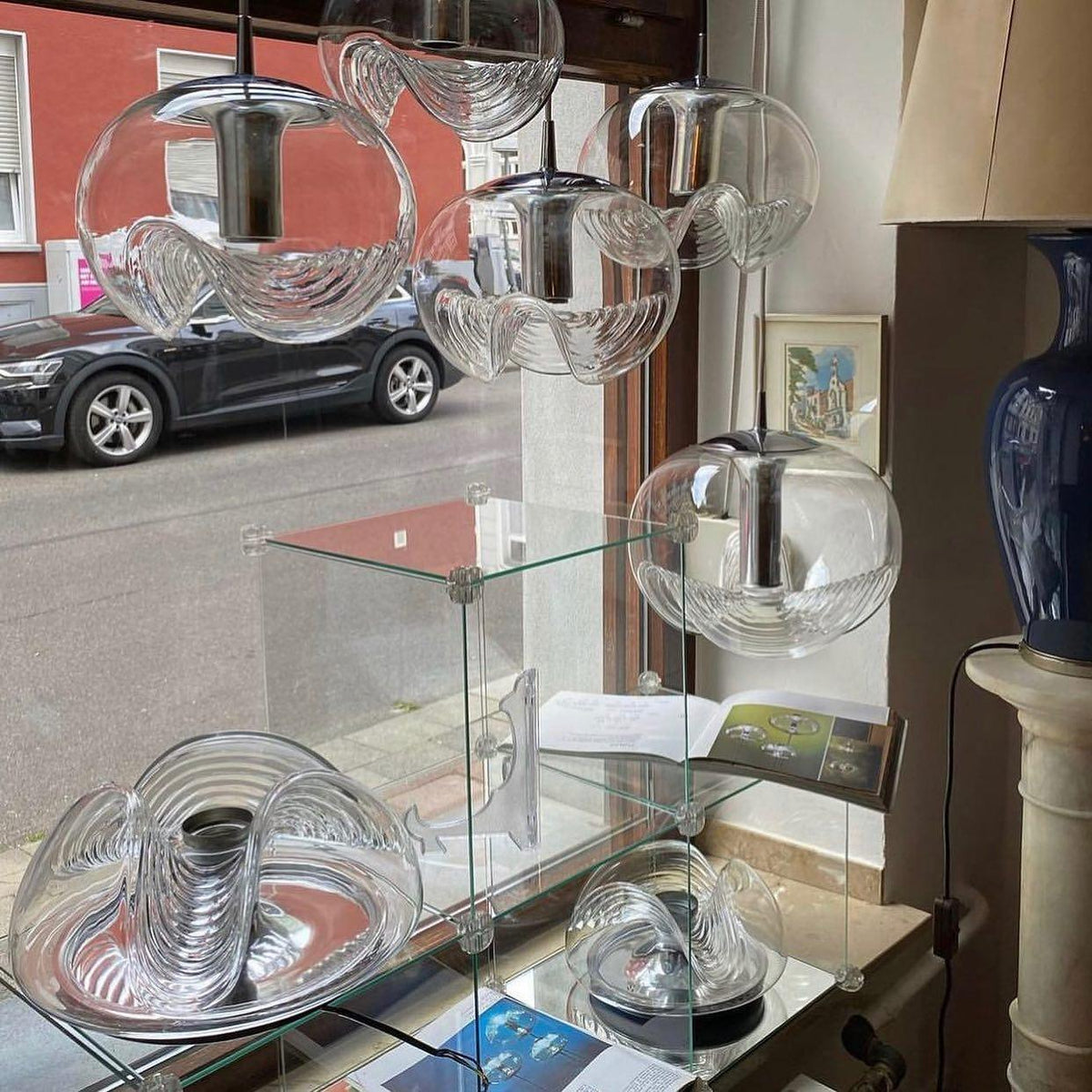 Displayed on a glass shelf in a shop window along a city street, this Clérachic pendant's clear glass shades sit beside parked cars visible outside.