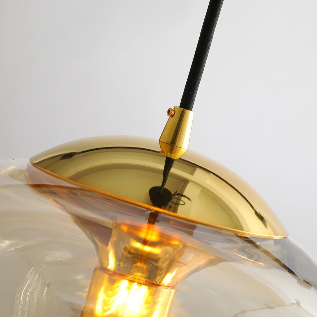 Close view of this Clérachic pendant's gold canopy and drop cable, its amber glass shade glowing warmly from the fitter below.