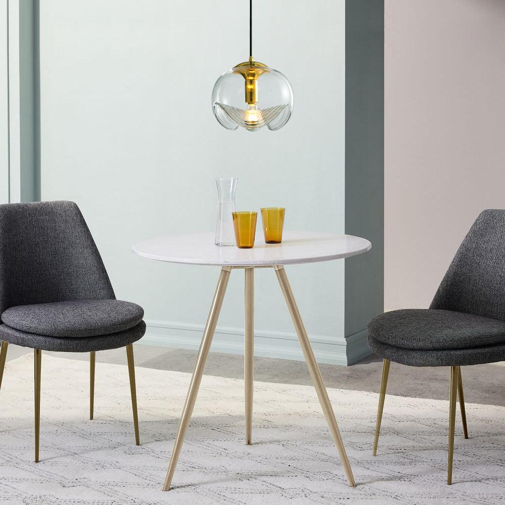 Above a round white dining table with amber glassware, this Clérachic pendant's smoky-toned glass shade and gold frame hang between two dark upholstered chairs.