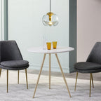 Above a round white dining table with amber glassware, this Clérachic pendant's smoky-toned glass shade and gold frame hang between two dark upholstered chairs.