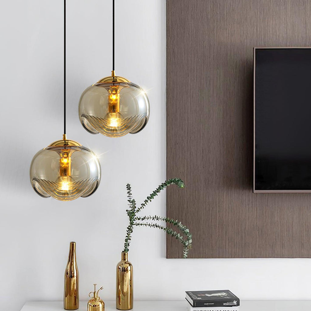 Two amber glass pendants with gold frames hang near a wall-mounted television, this Clérachic fixture paired above small gold vases on a console.