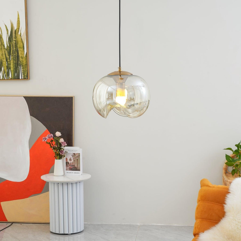Beside an abstract painting and a fluted white side table, this Clérachic pendant's clear glass shade and silver frame glow softly against a plain wall.