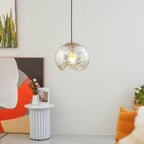 Beside an abstract painting and a fluted white side table, this Clérachic pendant's clear glass shade and silver frame glow softly against a plain wall.