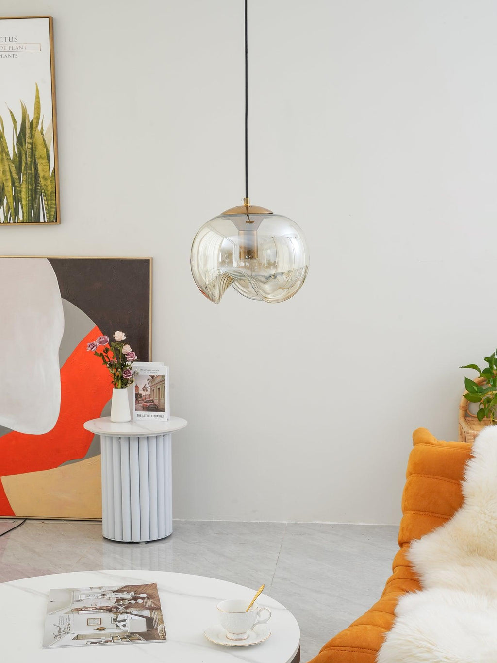 This Clérachic pendant's clear glass shade and silver frame hang above a fluted side table with fresh flowers and an orange throw pillow.