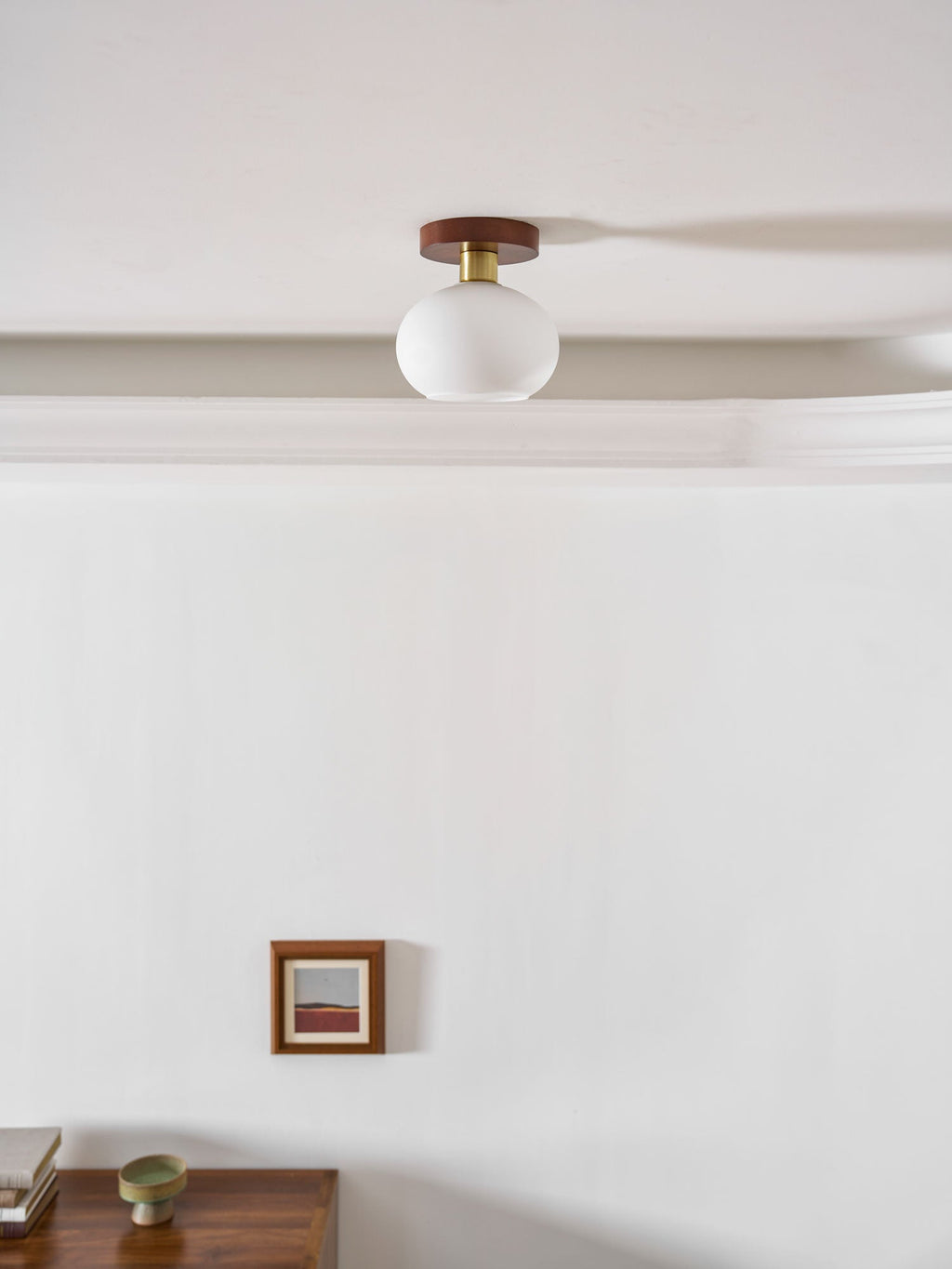 A nightstand and framed print sit below this Clérachic ceiling light, glowing warm along a crown-moulded ceiling.