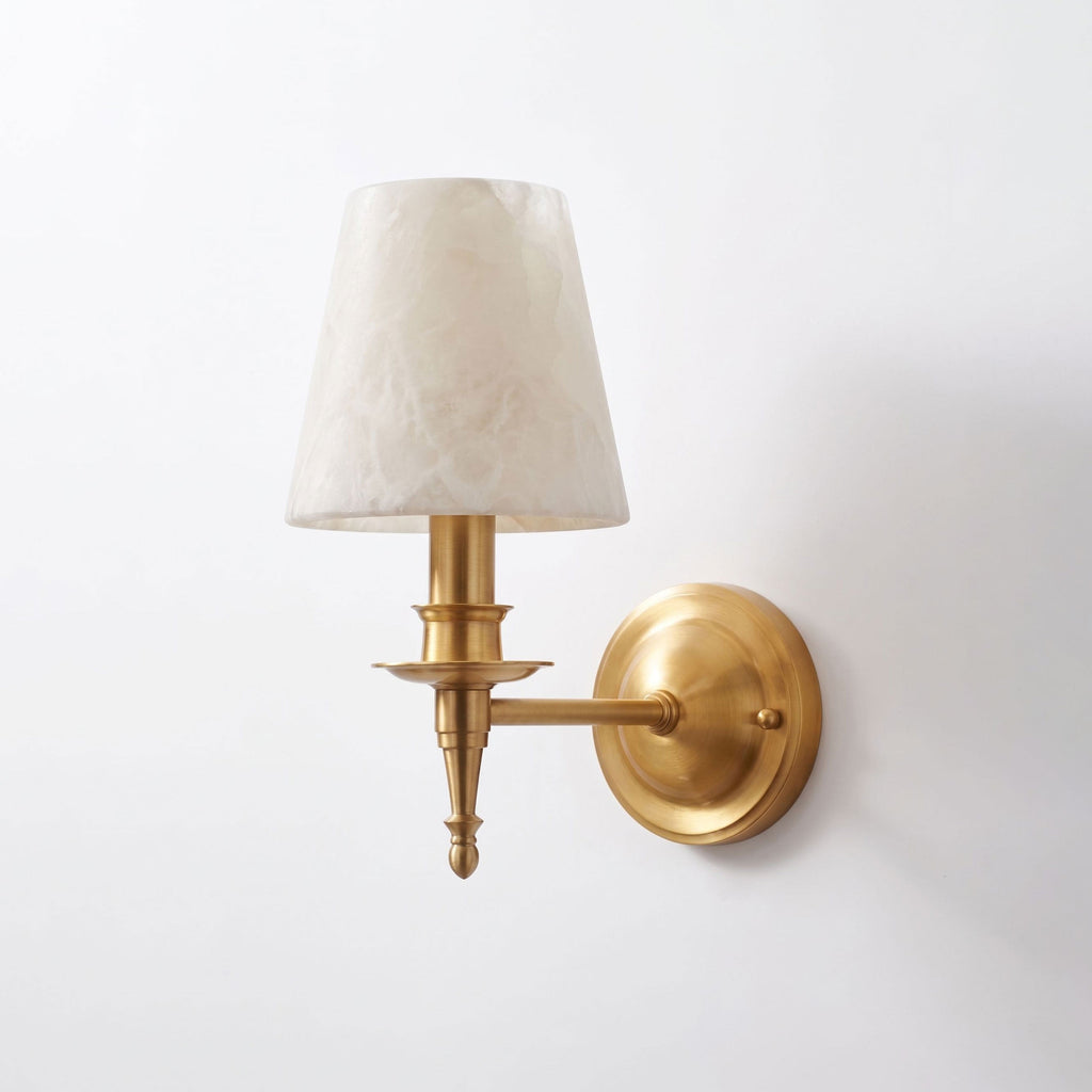 This brushed brass Clérachic wall sconce stands unlit against white, its alabaster cone shade catching soft light.