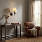 A leather armchair sits near this matte black Clérachic wall sconce, glowing above a wooden console styled with a vase.