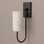 This Clérachic sconce in matte black stands lit on a plain taupe backdrop, its cylindrical alabaster shade glowing softly.
