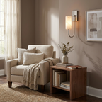 Beside a curtained window, a nickel Clérachic sconce glows above an armchair and a side table stacked with books.