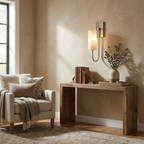 A nickel Clérachic sconce lights the wall above a console table stacked with books beside an upholstered chair.