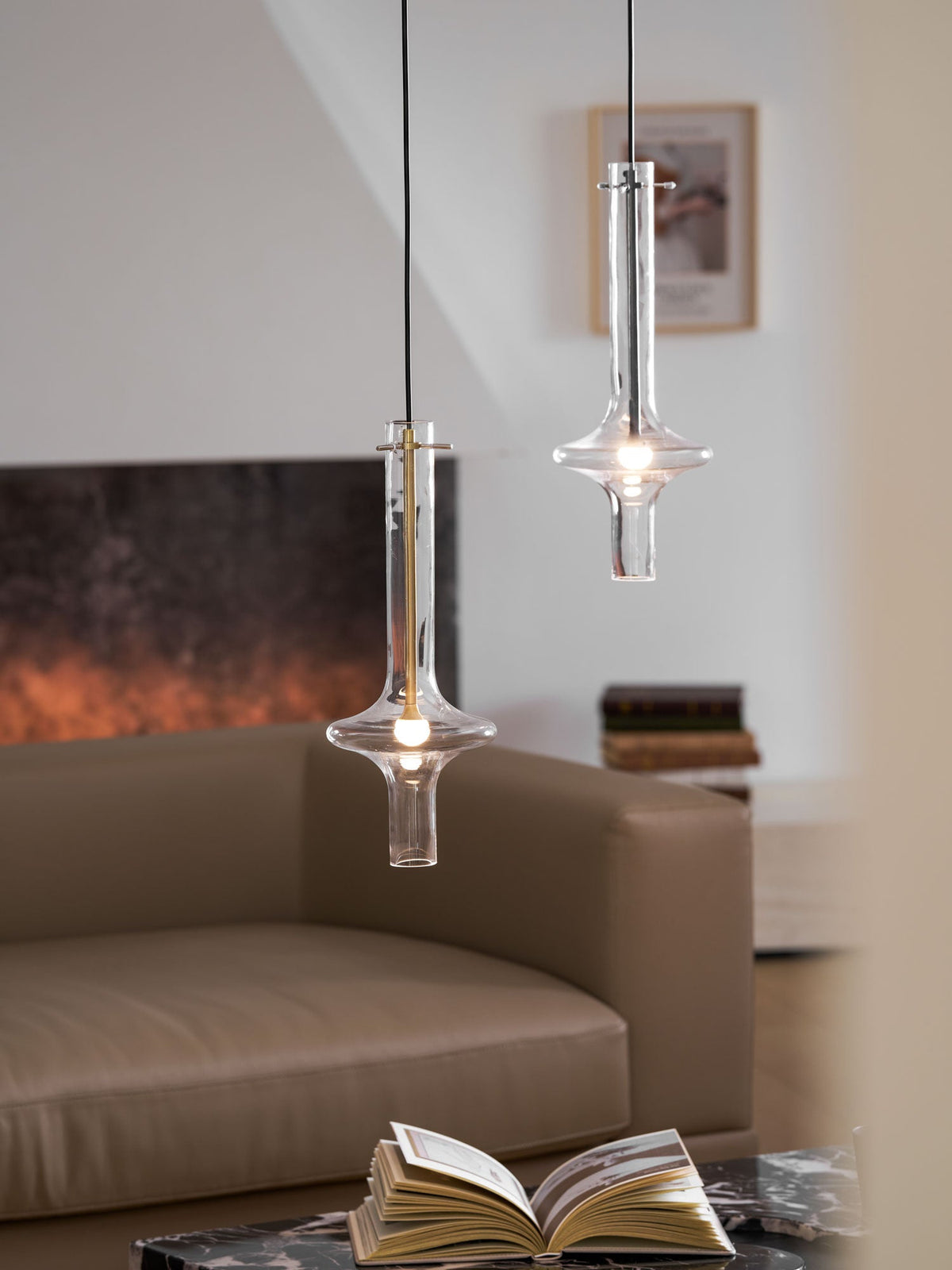 Two lit Clérachic pendants, one gold-stemmed and one clear, hang close together before a blurred living room with a tan sofa.