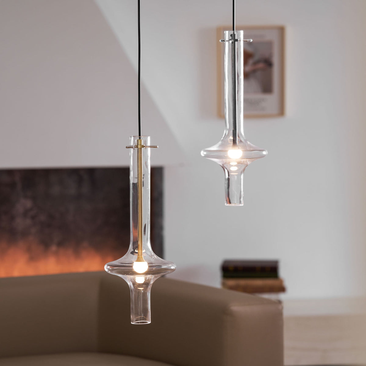 A gold-stemmed Clérachic pendant hangs closest to camera, its trumpet base lit, beside a taller clear pendant further back.