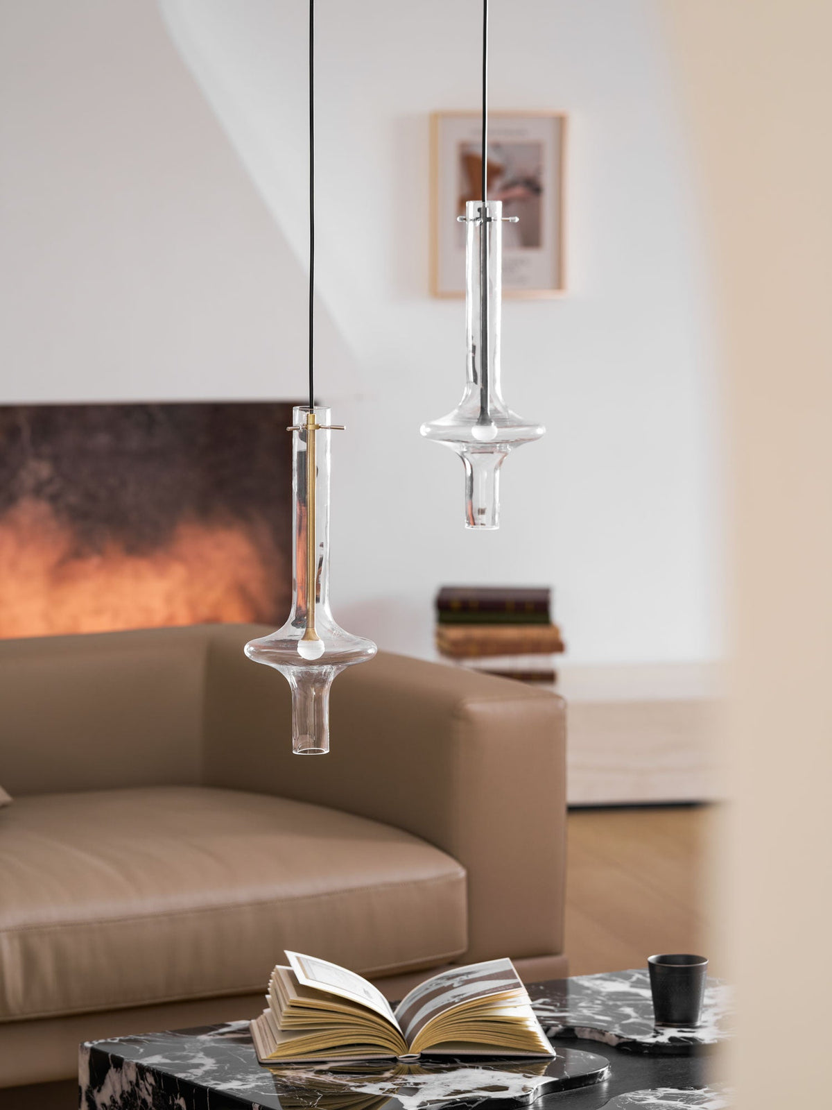 Two lit Clérachic pendants hang at different heights in a wider view of a living room, with a framed print on the wall behind.