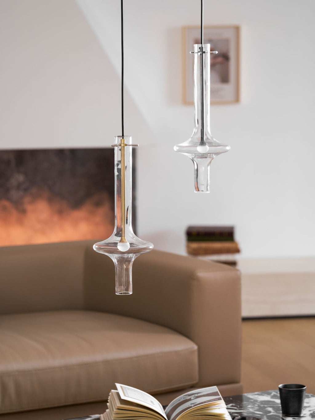A gold-stemmed Clérachic pendant hangs lower and to the left of a taller clear one, both lit before a blurred sofa and fireplace.