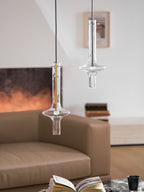 A gold-stemmed Clérachic pendant hangs lower and to the left of a taller clear one, both lit before a blurred sofa and fireplace.