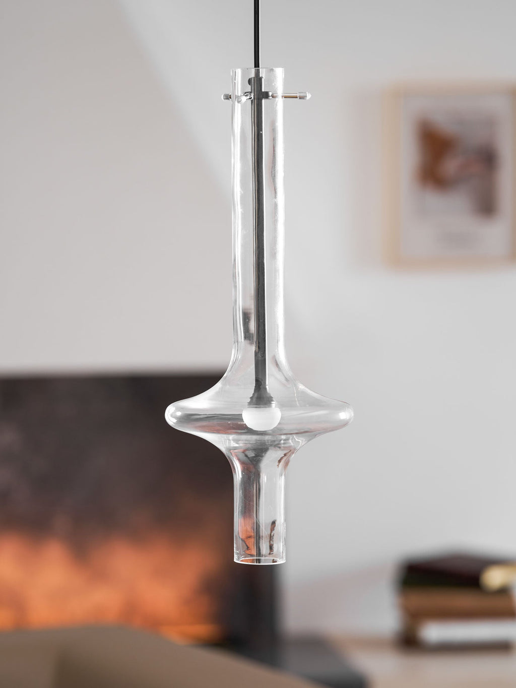 A single clear-glass Clérachic pendant hangs unlit in close view, with a blurred fireplace and sofa visible behind it.