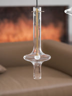 A close view of a lit clear-glass Clérachic pendant with a gold neck bar hangs before a blurred fireplace and tan sofa.