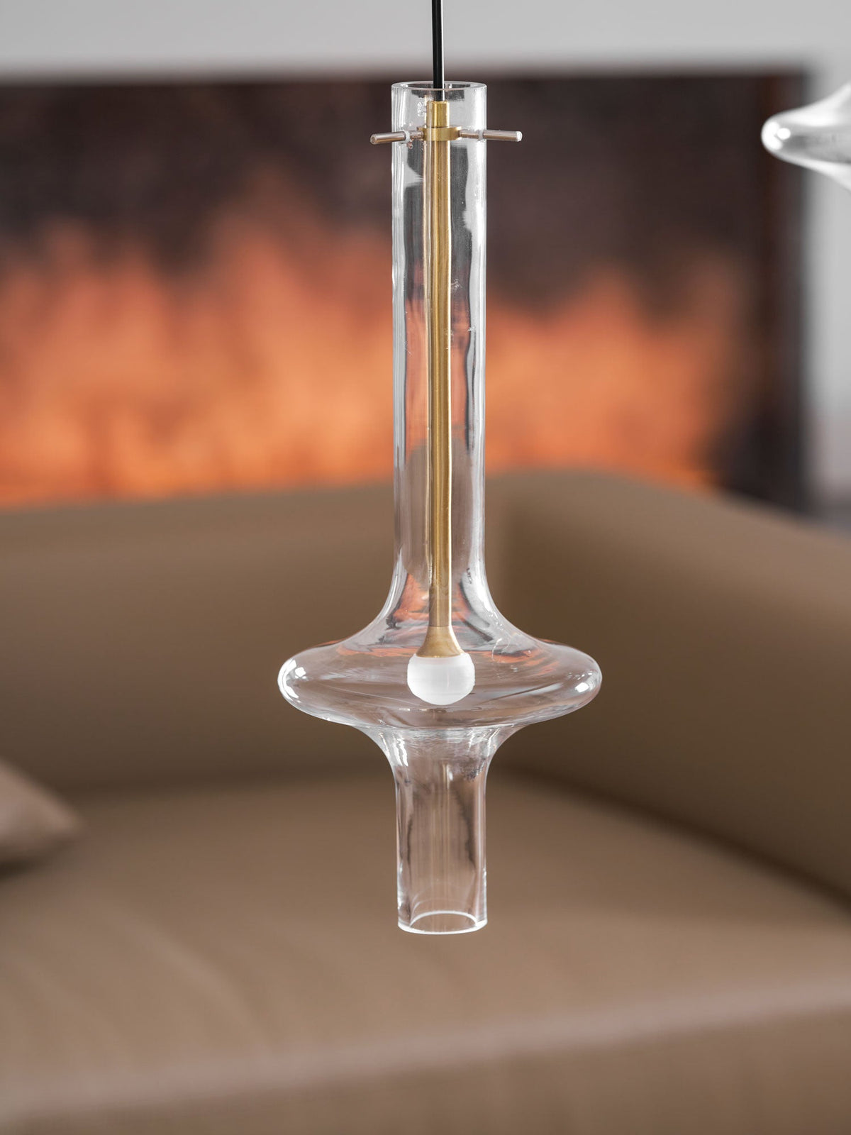A close view of a lit clear-glass Clérachic pendant with a gold neck bar hangs before a blurred fireplace and tan sofa.