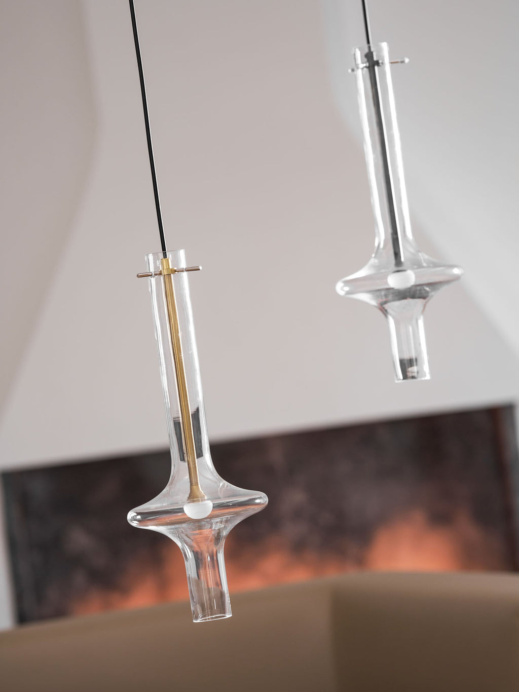 A lit gold-stemmed Clérachic pendant hangs lower, beside a taller clear pendant, before a blurred living room with a framed print.