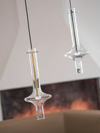 A lit gold-stemmed Clérachic pendant hangs lower, beside a taller clear pendant, before a blurred living room with a framed print.