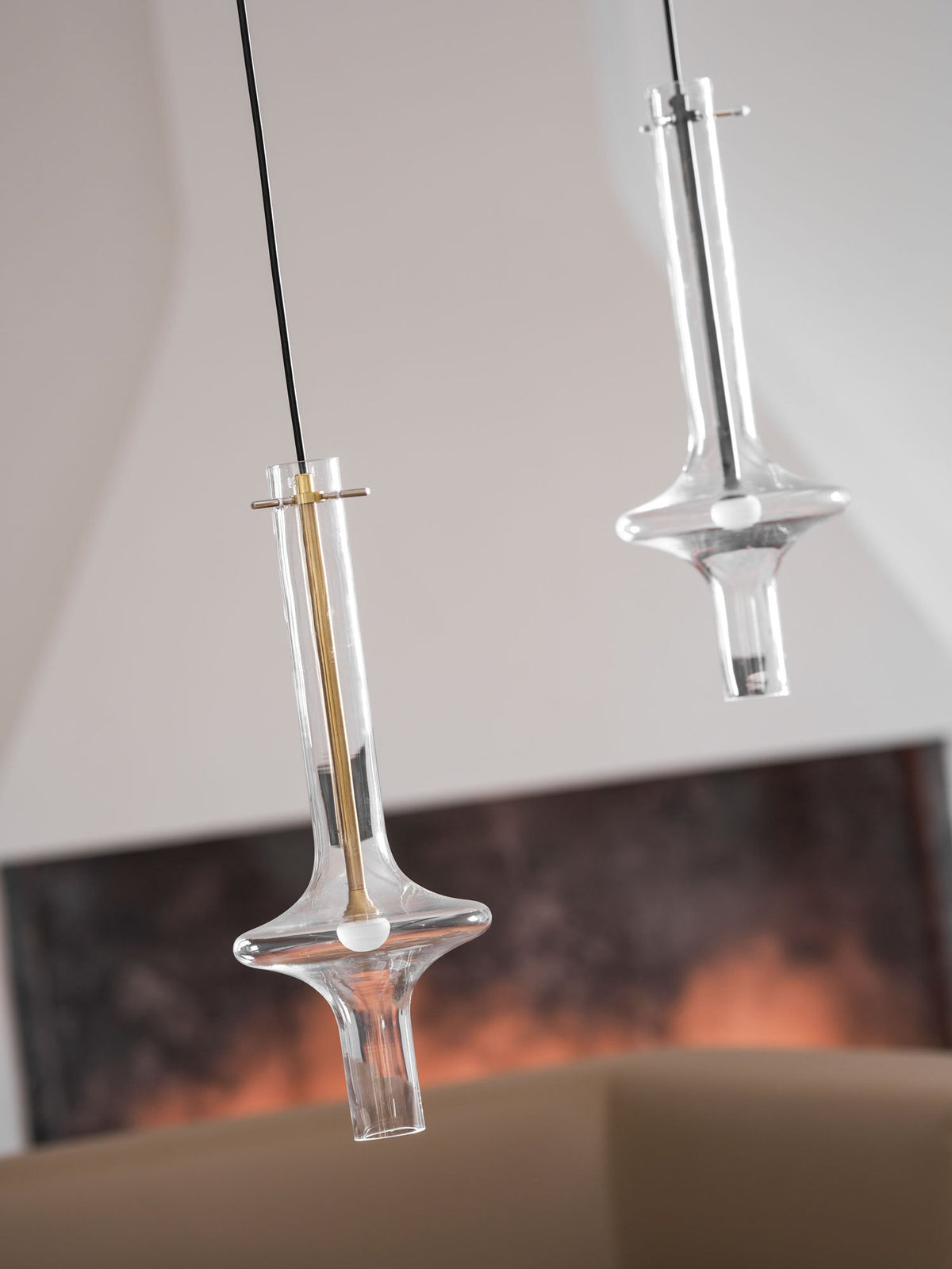 A lit gold-stemmed Clérachic pendant hangs lower, beside a taller clear pendant, before a blurred living room with a framed print.
