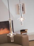 A lower gold-stemmed Clérachic pendant and a taller clear-stemmed one hang together, lit, before a blurred fireplace and framed print.