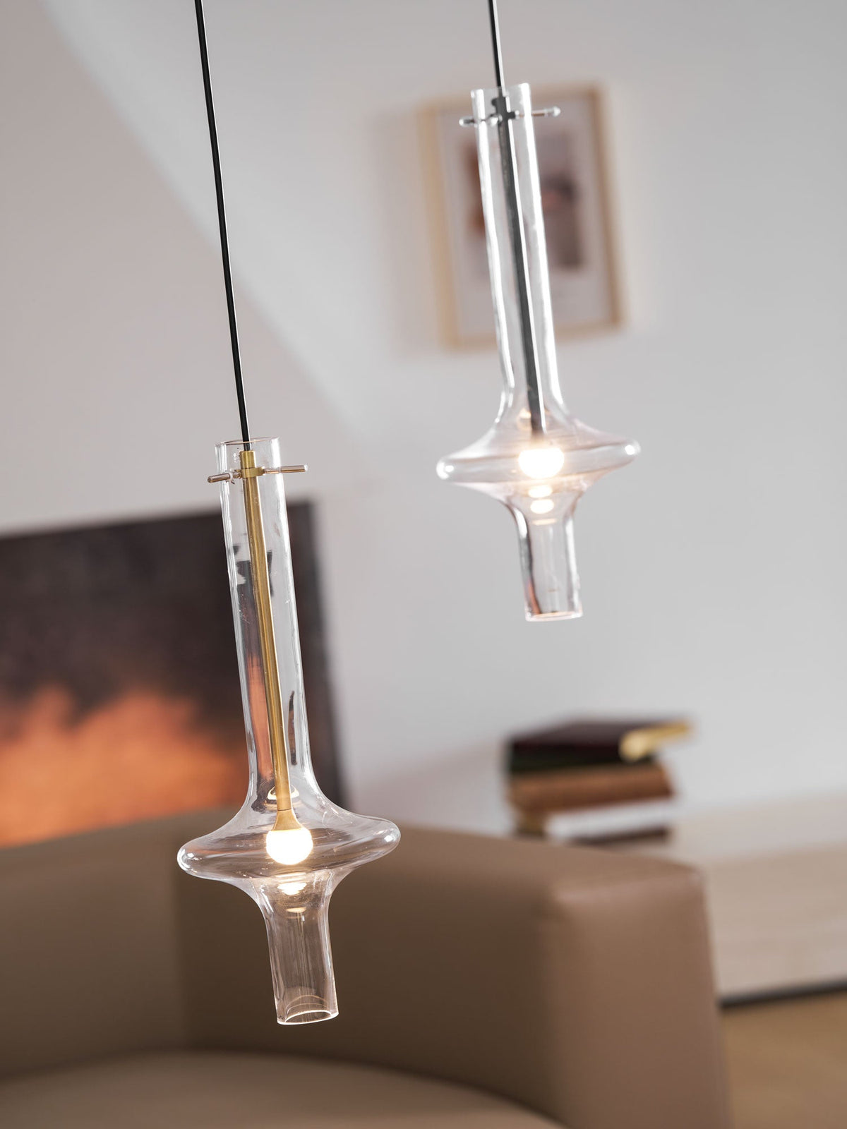 A lower gold-stemmed Clérachic pendant and a taller clear-stemmed one hang together, lit, before a blurred fireplace and framed print.