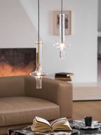 Two lit Clérachic pendants, gold and clear-stemmed, hang at staggered heights before a blurred living room with books on a table.