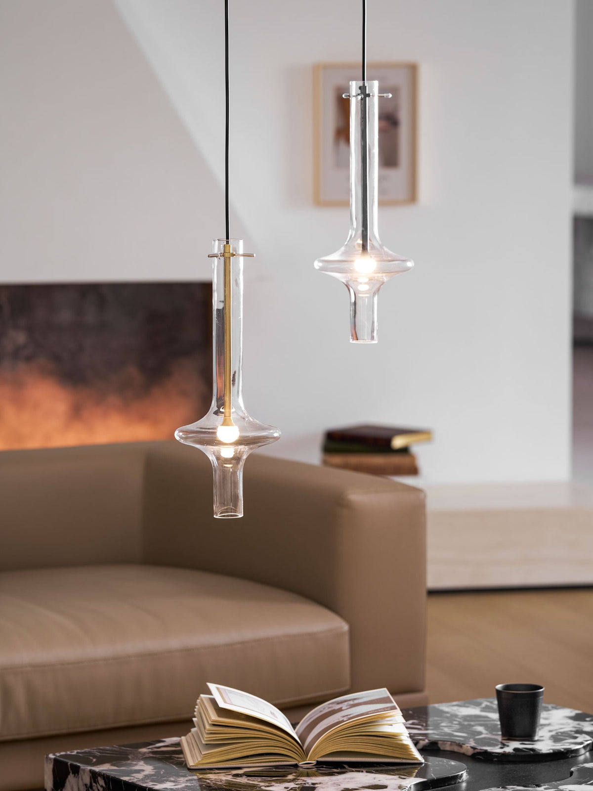 Two lit Clérachic pendants, gold and clear-stemmed, hang at staggered heights before a blurred living room with books on a table.