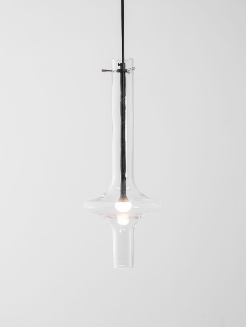 A single dark-stemmed Clérachic pendant with a gold crossbar glows in clear glass against a plain white backdrop.