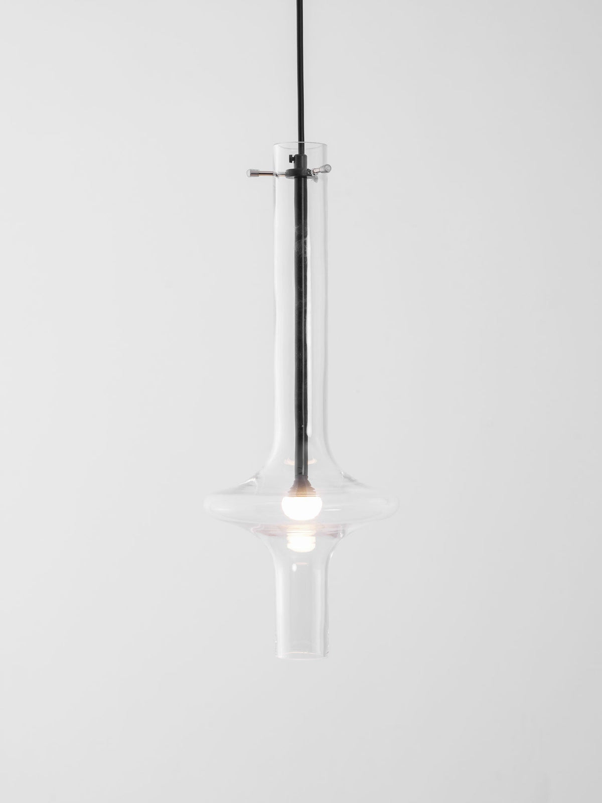 A single dark-stemmed Clérachic pendant with a gold crossbar glows in clear glass against a plain white backdrop.
