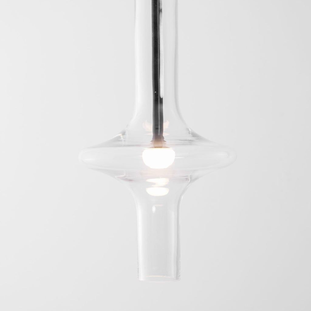 A close view shows the lit bulb inside this Clérachic pendant's flared clear glass base, with its gold crossbar visible above.