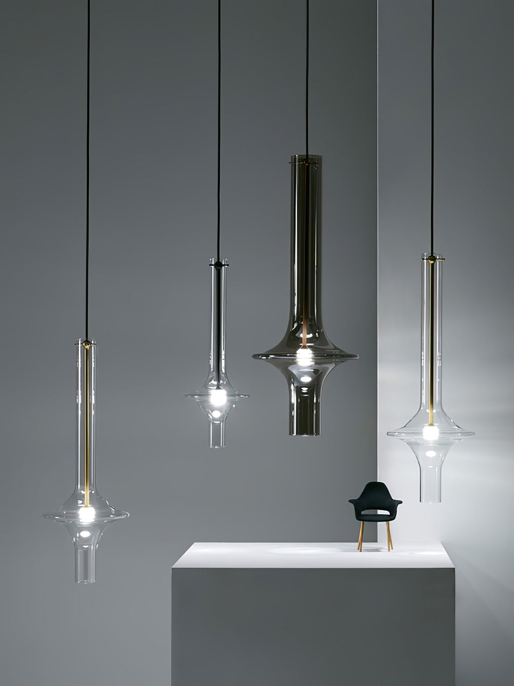 Four single Clérachic pendants in gold, black and clear finishes hang at different heights beside a plinth, against a blue-grey wall.