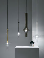 Four single Clérachic pendants in gold, black and clear finishes hang at different heights beside a plinth, against a blue-grey wall.