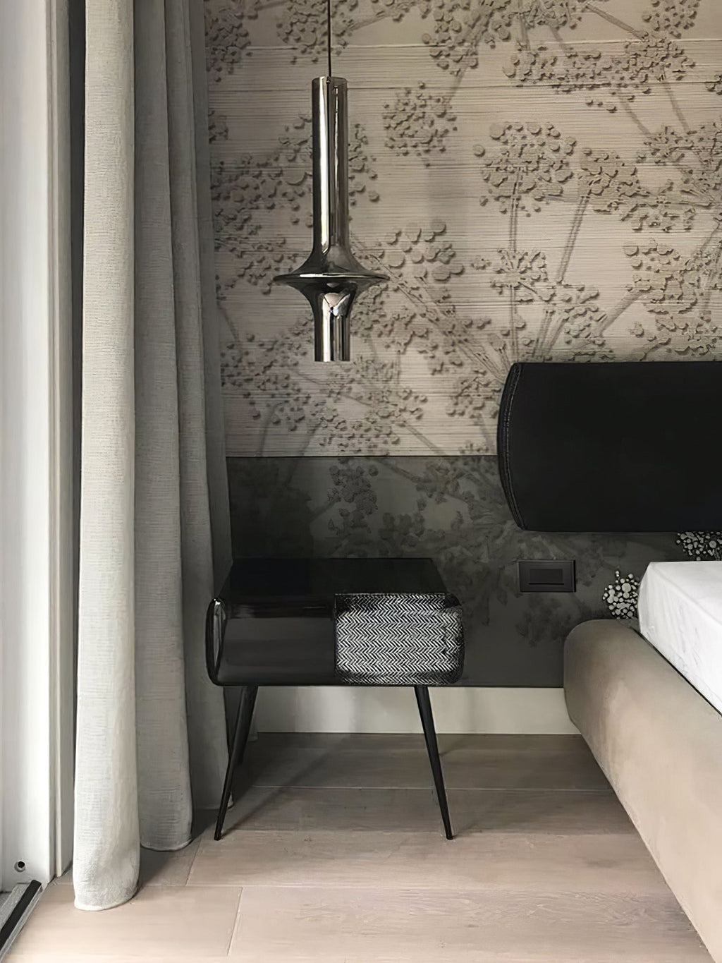 A single nickel-toned Clérachic pendant hangs beside a bed with textured floral wallpaper and a dark bedside table.