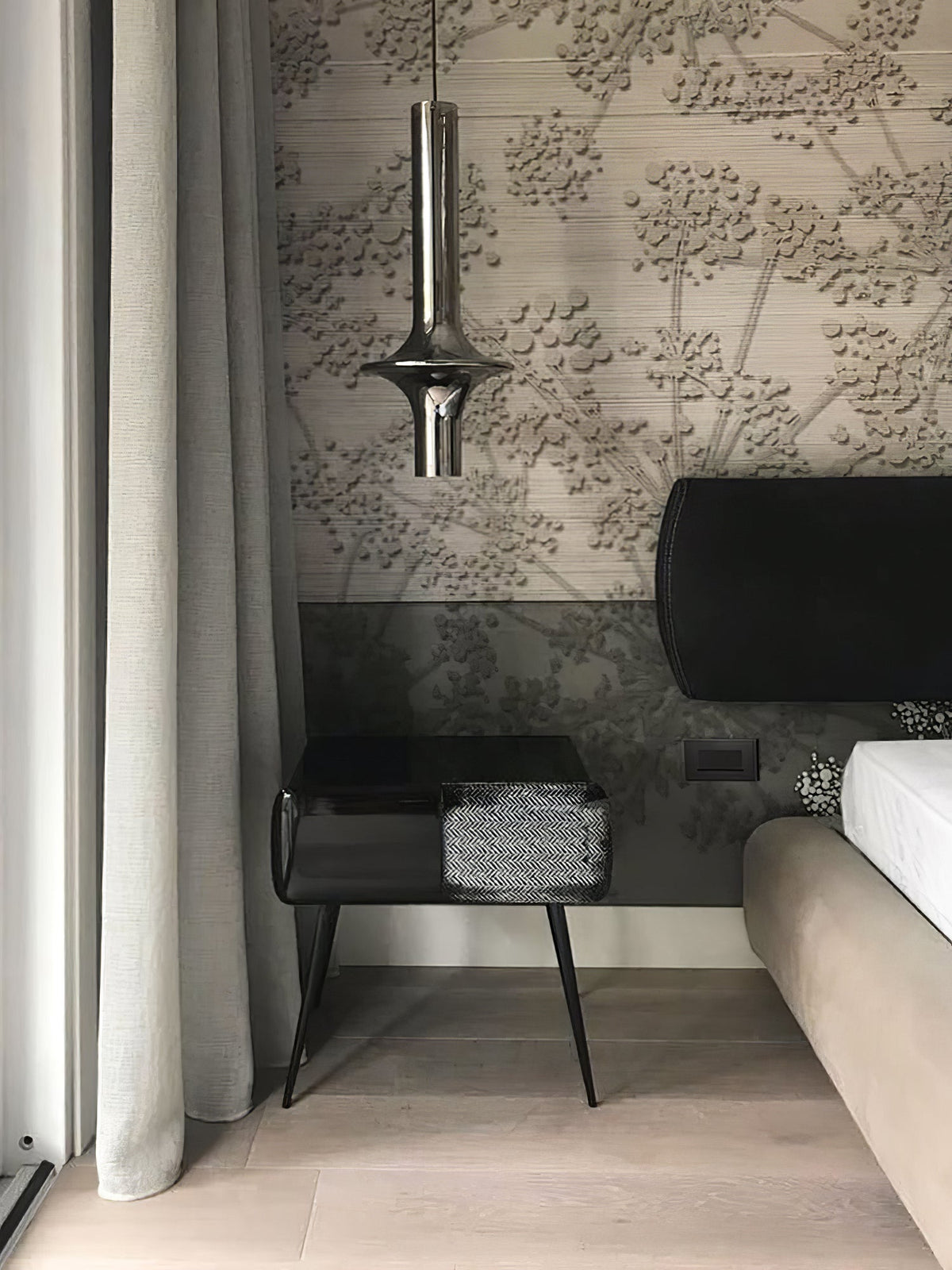 A single nickel-toned Clérachic pendant hangs beside a bed with textured floral wallpaper and a dark bedside table.
