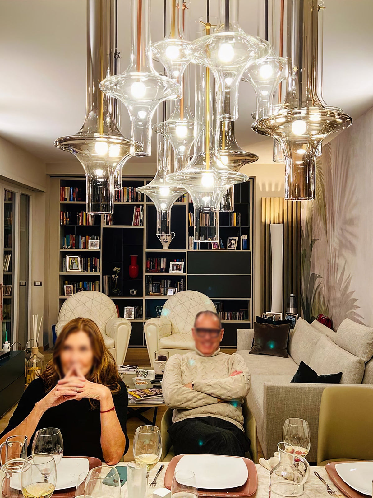A large cluster of lit Clérachic pendants hangs above a dining table set for two, with a bookshelf filling the background.