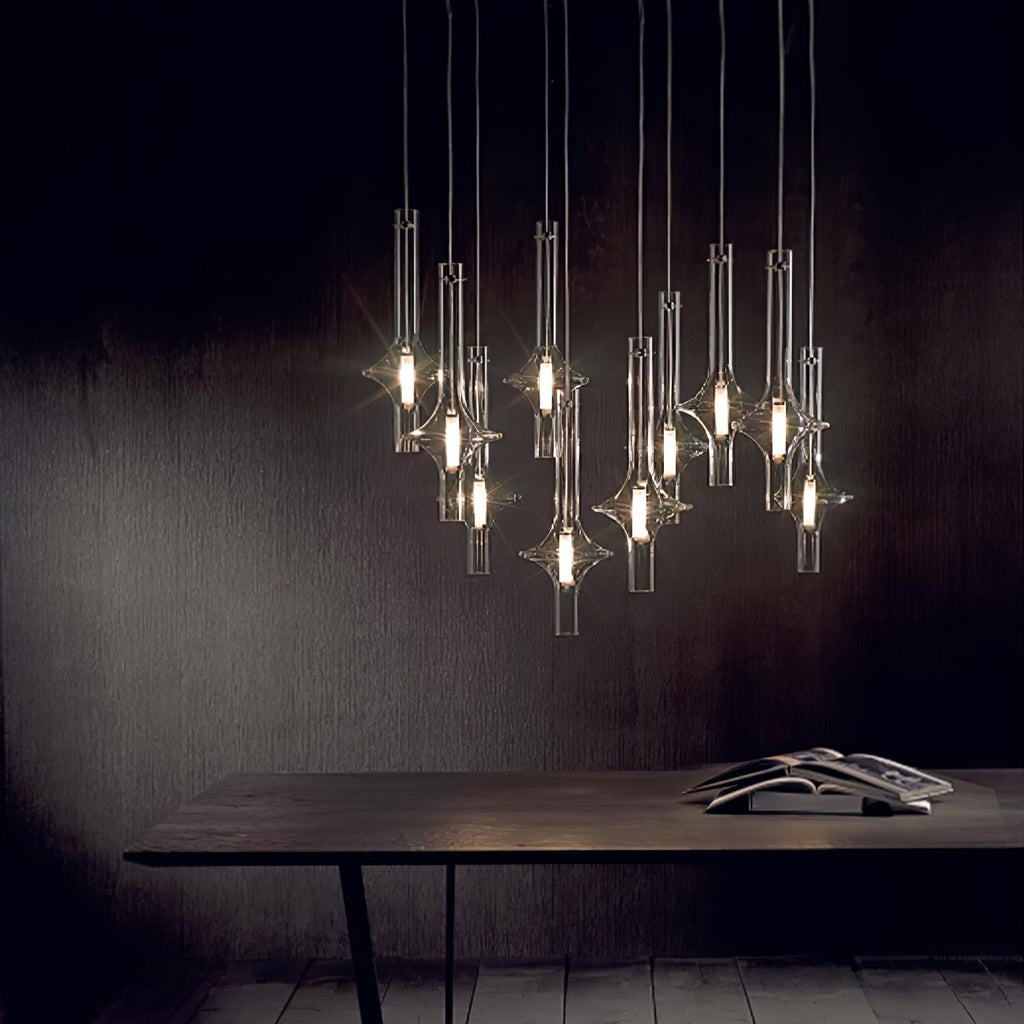 A tall cluster of lit Clérachic pendants hangs over a dark wood dining table stacked with books, against a black studio wall.