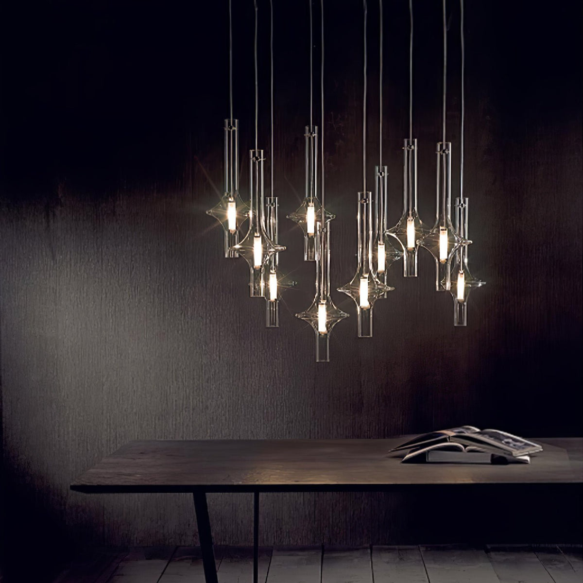 A tall cluster of lit Clérachic pendants hangs over a dark wood dining table stacked with books, against a black studio wall.
