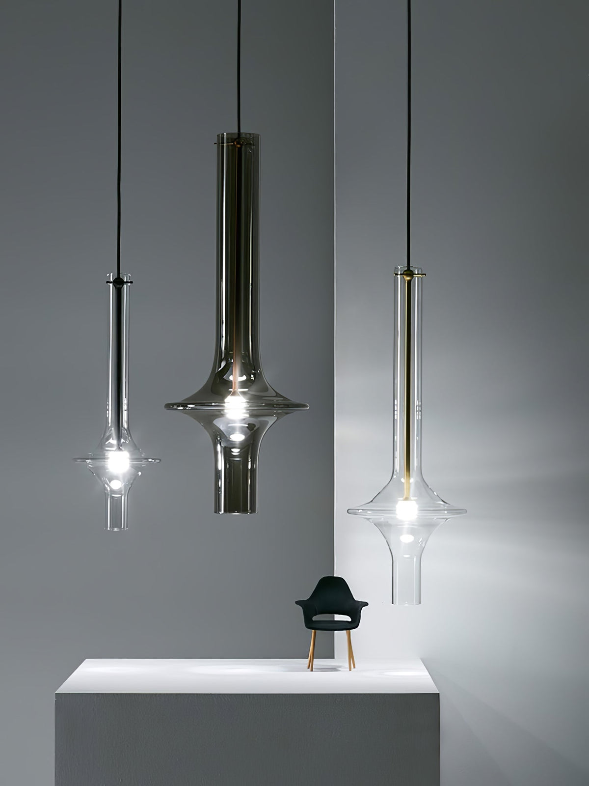 Three single Clérachic pendants in black, smoky and clear glass hang at staggered heights beside a plinth and a small chair figurine.